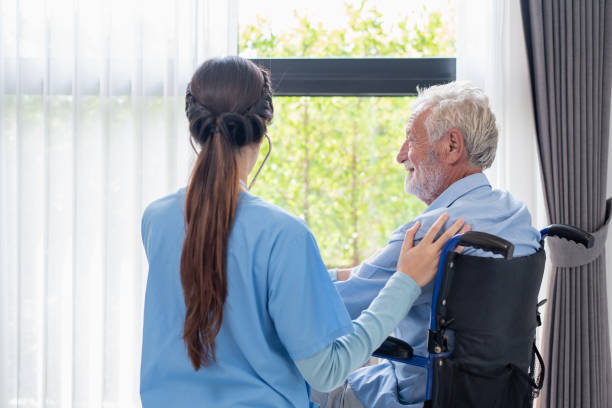 A nurse and a senior white man look out the window with a smile.